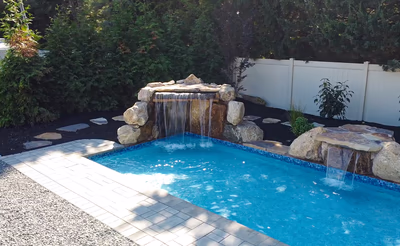 Kapsuriat C - Williamstown, NJ - This custom installation features a multi-tier natural stone waterfall cascading into a gunite pool, surrounded by large boulders and a white picket fence. The pool has a clean, blue finish with a tiled edge, and the surrounding area is landscaped with gravel and additional rocks for a natural, serene look. The craftsmanship is evident in the seamless integration of the waterfall and pool, creating a tranquil outdoor living space in Williamstown, NJ.