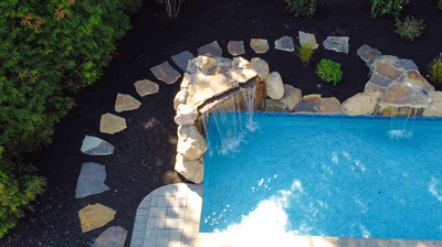 Kapsuriat D - Williamstown, NJ - This custom installation features a natural stone waterfall cascading into a gunite pool, surrounded by strategically placed boulders and dark mulch. The waterfall is constructed with carefully stacked irregular stones creating a multi-tiered cascade that flows into the deep blue pool below. The surrounding area is landscaped with low shrubs and additional boulders, enhancing the natural grotto aesthetic of this Williamstown, NJ backyard oasis.