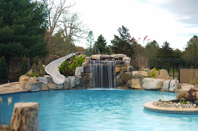 Williams A - Port Republic, NJ - This custom installation features a multi-tier natural stone waterfall cascading into a gunite pool, with integrated rock formations creating a grotto-like environment. The waterfall includes a smooth concrete slide embedded in the stone structure, allowing for both visual appeal and recreational use. Surrounded by mature landscaping and a black metal fence, the project demonstrates exceptional craftsmanship through seamless integration of water features, natural materials, and functional design elements.