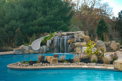 Williams B - Port Republic, NJ - This custom installation features a multi-tier natural stone waterfall cascading into a gunite pool, with integrated rock formations creating a grotto-like environment. The waterfall includes a smooth, curved slide embedded in the stone structure, allowing for both visual and functional appeal. Surrounded by carefully placed boulders and lush landscaping, the project demonstrates exceptional craftsmanship and attention to detail in creating a serene backyard oasis.