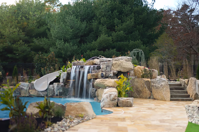 Williams C - Port Republic, NJ - This custom installation features a multi-tier natural stone waterfall cascading into a gunite pool, with large river rocks forming the cascade structure. The design incorporates a smooth concrete slide integrated into the left side of the waterfall, creating a family-friendly water feature. The surrounding area includes a paved patio with stone accents, lush landscaping with shrubs and flowering plants, and a black metal fence providing privacy in a serene backyard setting.