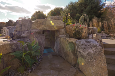 Williams E - Port Republic, NJ - This custom outdoor installation features a natural stone waterfall cascading into a small, inviting pool. The waterfall is constructed from large, irregularly shaped boulders that create a rustic, organic feel, with water flowing smoothly over the stone surface. The pool area is bordered by additional boulders and lush greenery, enhancing the natural aesthetic. The setting is part of a well-maintained backyard with a small shed visible in the background, creating a serene and tranquil environment.