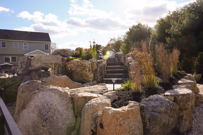 Williams F - Port Republic, NJ - This custom outdoor living project features a natural stone waterfall cascading over large boulders into a rocky pool area. The waterfall is constructed with carefully placed natural stone, creating a multi-tiered cascade that flows seamlessly into a textured pool bed. The surrounding landscaping includes tall grasses and shrubs, enhancing the natural, serene atmosphere of the Port Republic, NJ backyard setting.