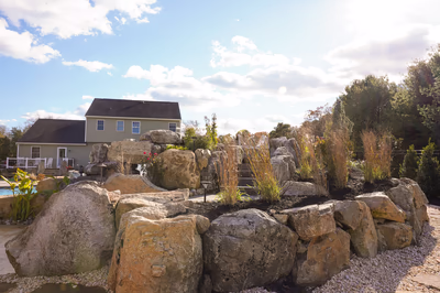 Williams G - Port Republic, NJ - This professional installation features a multi-tier natural stone waterfall cascading into a gunite pool, surrounded by large, irregularly shaped boulders that create a rugged, organic aesthetic. The waterfall includes a tiered cascade with a spillway leading into the pool, showcasing high-quality craftsmanship and attention to detail. The surrounding landscape incorporates tall grasses, shrubs, and a well-maintained lawn, enhancing the natural, serene setting of this outdoor living space in Port Republic, NJ.