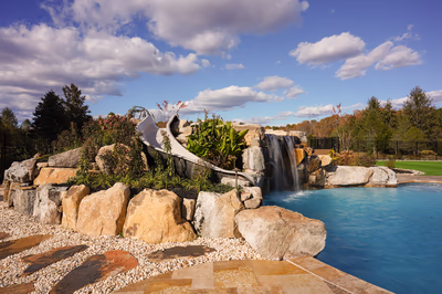 Williams H - Port Republic, NJ - This custom installation features a multi-tier natural stone waterfall cascading into a gunite pool, accented with large river boulders and integrated slide. The craftsmanship showcases seamless integration of water features with surrounding landscaping, creating a serene backyard oasis under a bright blue sky.