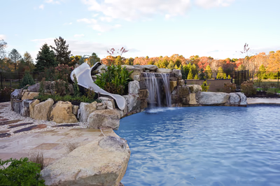 Williams I - Port Republic, NJ - This custom outdoor living project features a natural stone waterfall cascading into a gunite pool, with integrated slide embedded in the stonework. The waterfall includes multiple tiers of rough-hewn boulders creating a multi-level cascade, while the pool showcases clean lines and a smooth surface. The surrounding area is landscaped with seasonal foliage and a black metal fence, creating a serene backyard retreat in Port Republic, NJ.