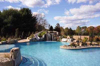 Williams K - Port Republic, NJ - This custom installation features a natural stone waterfall cascading over layered boulders into a pristine gunite pool. The design incorporates a smooth concrete slide integrated into the waterfall structure, allowing swimmers direct access from the waterfall into the pool. The surrounding area is landscaped with additional boulders and low-lying vegetation, creating a seamless transition between the water feature and the surrounding backyard environment.