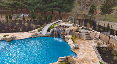 Williams L - Port Republic, NJ - This custom installation features a natural stone waterfall cascading from a tiered rock formation into a gunite pool. The waterfall includes a smooth concrete slide integrated into the rock structure, allowing access directly into the pool. The surrounding area is landscaped with carefully placed boulders, stepping stones, and low-maintenance shrubs, creating a serene backyard oasis in Port Republic, NJ.