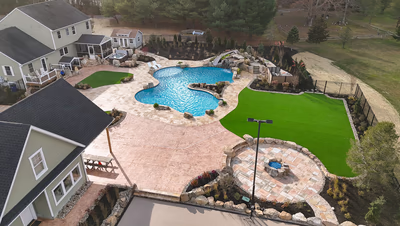 Williams N - Port Republic, NJ - This professionally installed outdoor living space features a multi-tier natural stone waterfall cascading into a gunite pool with a freeform design. The waterfall incorporates large boulders and carefully placed stone accents that create a seamless transition between water features and surrounding landscaping. The project demonstrates exceptional craftsmanship through precise stonework, integrated lighting, and thoughtful integration with the existing backyard environment, including artificial turf, a fire pit area, and well-maintained garden beds.
