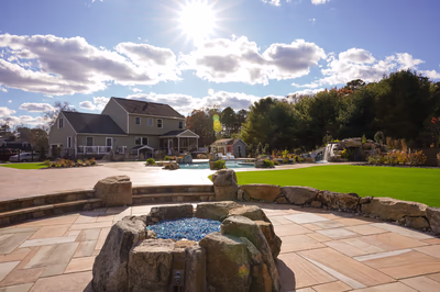 Williams Q - Port Republic, NJ - This professionally installed outdoor living project features a multi-tier natural stone waterfall cascading into a gunite pool, accented with large river boulders. The construction demonstrates exceptional craftsmanship with seamless integration of the waterfall into the pool deck, creating a tranquil water feature surrounded by meticulously landscaped greenery. The setting includes a spacious patio area with interlocking stone pavers and a well-maintained lawn, enhancing the luxurious backyard ambiance.