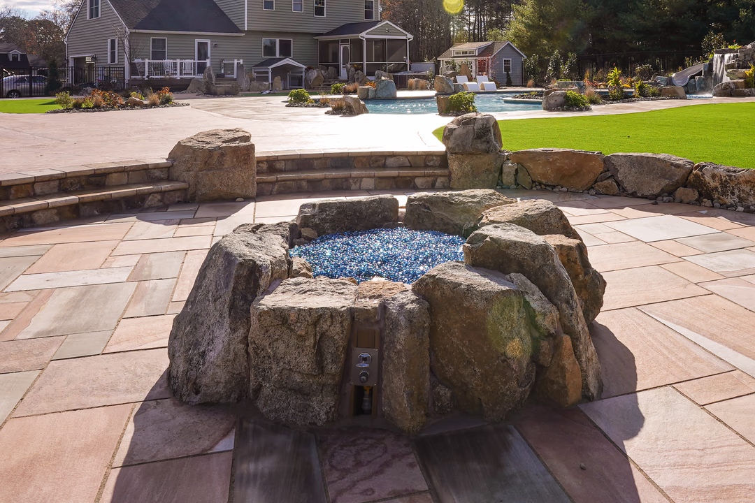 Williams R - Port Republic, NJ - This custom outdoor living project features a natural stone waterfall cascading over large boulders into a gunite pool. The waterfall includes a blue glass pebble bed at its base, creating a visually striking contrast against the rugged stone. The surrounding area is paved with large, light-colored stone tiles, and the design integrates seamlessly with the landscaped backyard, which includes a well-maintained lawn, additional rock formations, and a small pool area with a waterfall and slide.