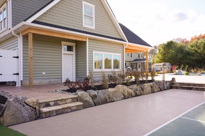 Williams S - Port Republic, NJ - This professional installation features a natural stone waterfall integrated into a landscaped patio area adjacent to a house. The waterfall uses large, irregularly shaped boulders arranged in a tiered cascade that flows into a small pool or water feature. The surrounding patio is constructed with clean concrete surfaces and brick pavers, creating a seamless transition between the house's wooden deck and the outdoor living space. The project demonstrates excellent craftsmanship with precise stone placement and thoughtful integration of hardscape elements, set in a well-maintained backyard with mature trees and additional landscaping features visible in the background.