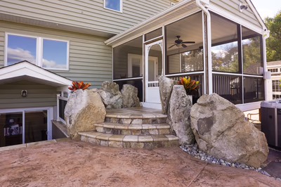 Williams U - Port Republic, NJ - This custom outdoor living project features a natural stone waterfall cascading down multi-tiered steps, integrated with large, rugged boulders for a rustic aesthetic. The waterfall is constructed with high-quality natural stone, creating a seamless flow that enhances the serene backyard setting. The surrounding area includes a spacious patio with a warm, textured concrete finish, complemented by lush greenery and a screened porch, providing both functionality and visual appeal.