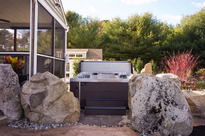 Williams W - Port Republic, NJ - This custom hot tub installation features a sleek, modern design with a dark wood frame and a clean, white cover. It is surrounded by large, irregularly shaped natural stones that create a rustic, inviting ambiance. The setting is enhanced by a well-maintained garden with lush greenery, providing a tranquil outdoor relaxation space adjacent to a screened porch.